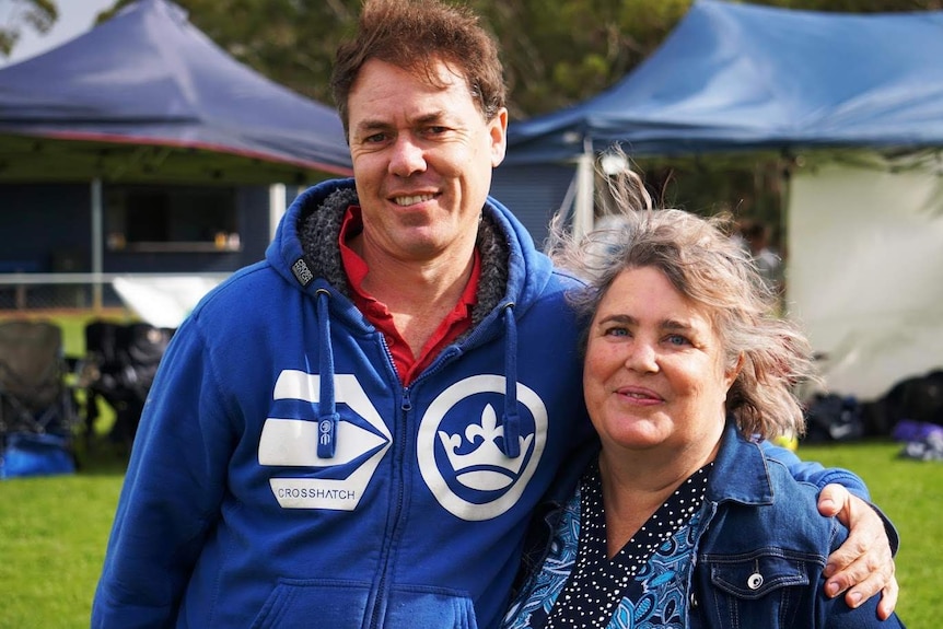 A man with a blue parker and a woman with a denim jacket stand smiling, arms around each other, in front of sport gazebos.