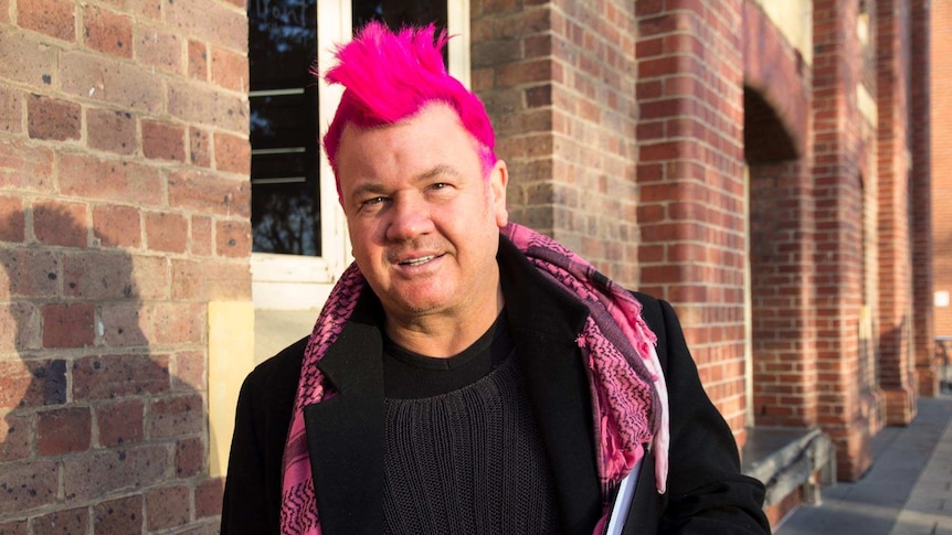 A man with spiky pink hair looks into the camera