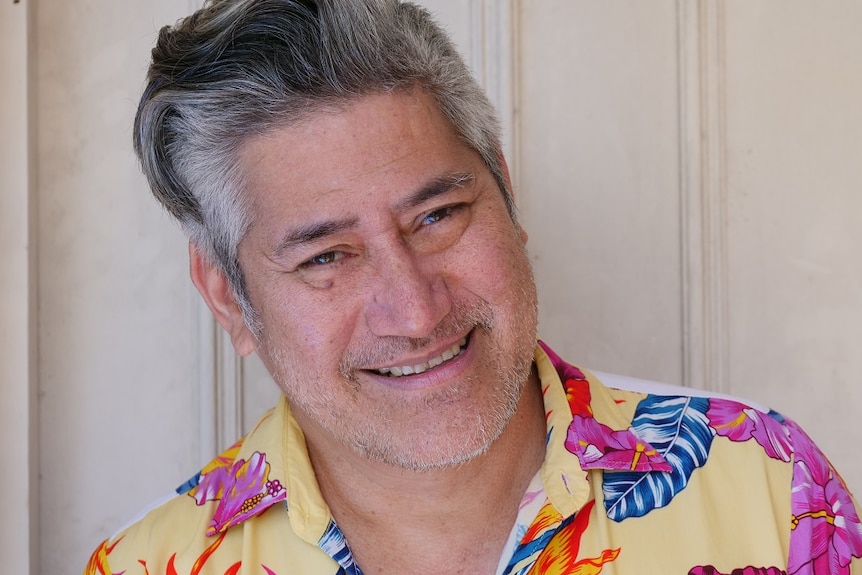 A man with black and grey hair smiling iin front of a door on a sunny day and wearing a floral shirt