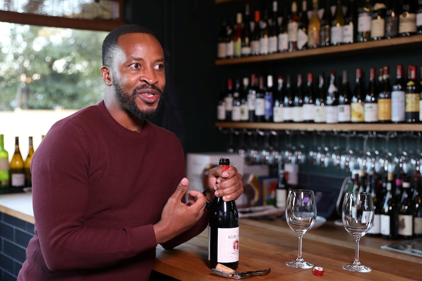 Tinashe Nyamudoka, with short black hair and red shirt, sits at a bar in mid-speech, holding a bottle of red wine.