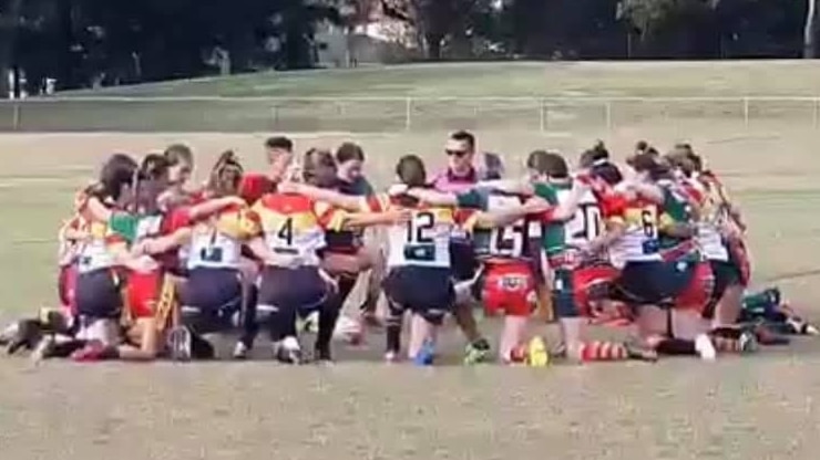 Female rugby league players kneeling in a circle in the middle of a field.