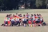 Female rugby league players kneeling in a circle in the middle of a field.