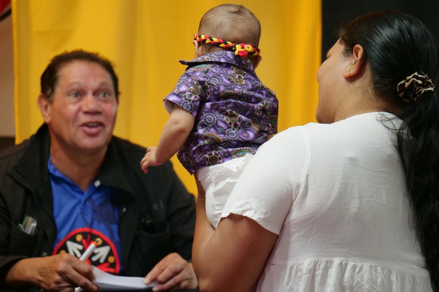 Jeffrey smiling at a young baby in a purple shirt being held up by her mum, yellow background.