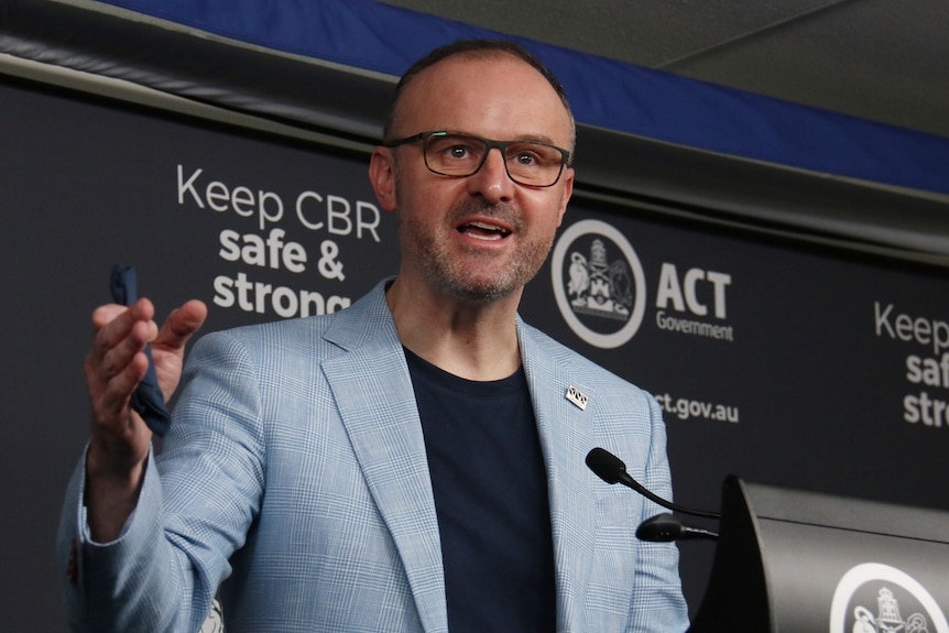 A man in a jacket at glasses speaks at a podium.