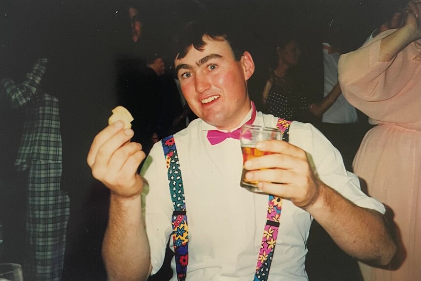 A man in suspenders and a pink bow tie holds up a beer.