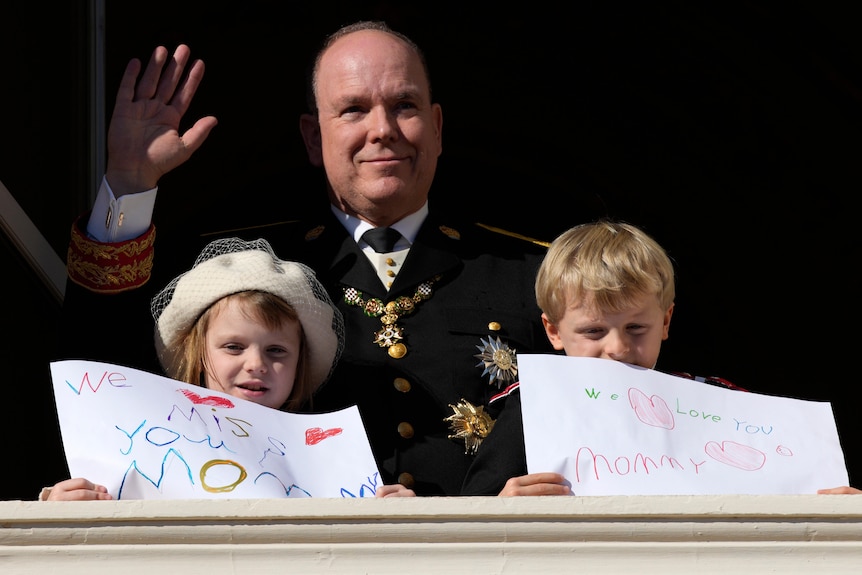 Monaco's Prince Albert in uniform waves from a balcony, with his children in front holding signs saying 'we miss mommy'