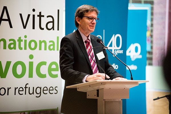 A man with brown hair and glasses speaking at a podium in front of banners.