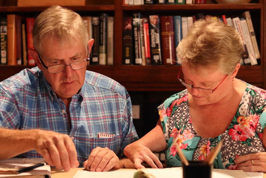 A man and a woman, both wearing glasses, look through documents. A shelf of books sits in the background