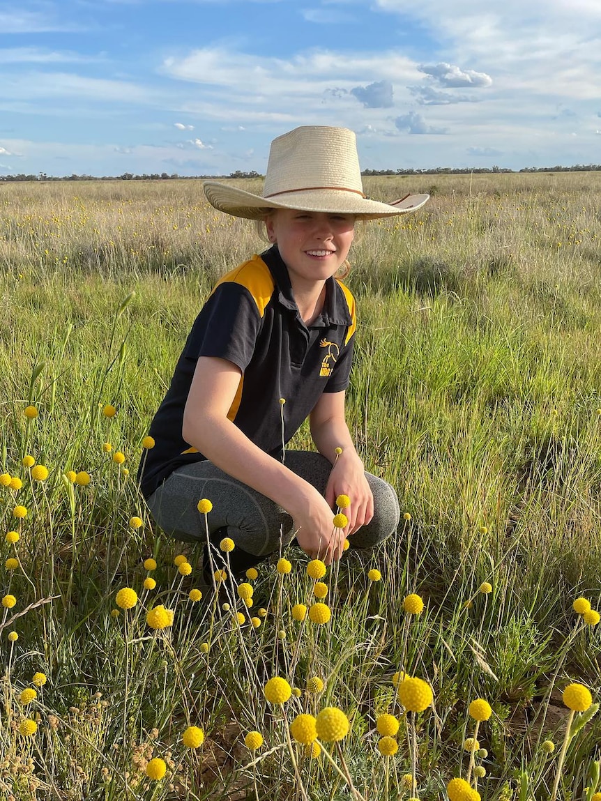 A girl in an akubra crouched in a paddock surrounded by yellow wildflowers