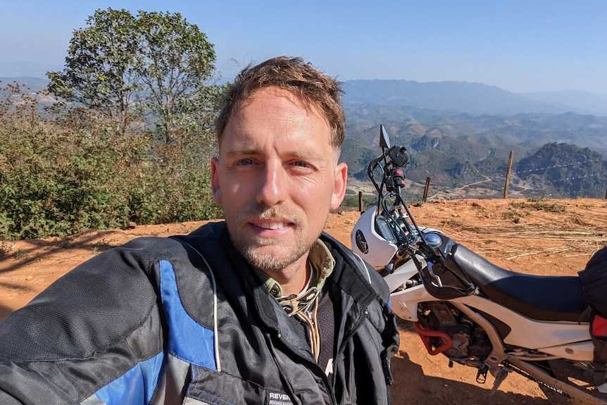 Young man with short brown hair and a black and blue motorcycle jacket stands with his motorbike and countryside in background.