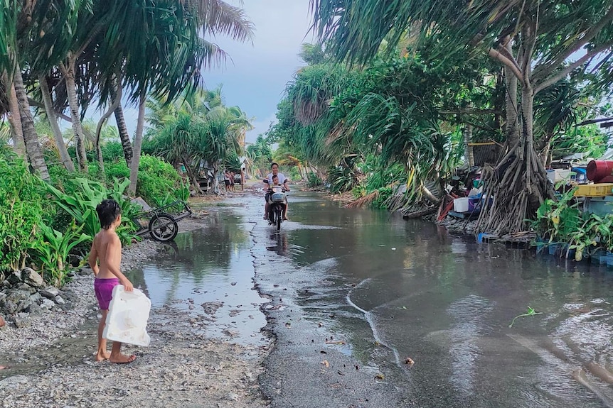 A child stands next to a flooded road while a mother rides a bike with her baby.