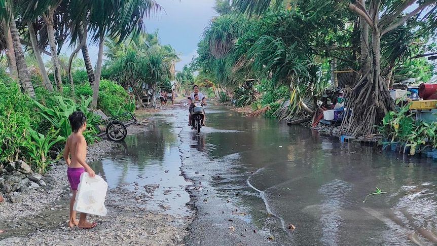 A child stands next to a flooded road while a mother rides a bike with her baby.