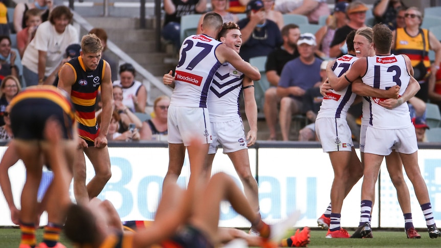 A group of AFL players throw their arms around each other in celebration as their opponents look dejected after a game.. 