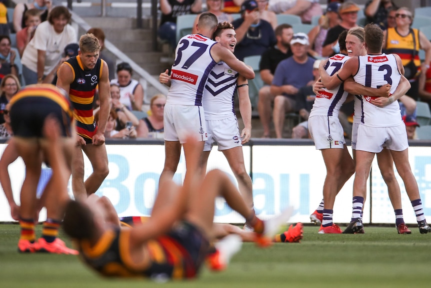 A group of AFL players throw their arms around each other in celebration as their opponents look dejected after a game.. 