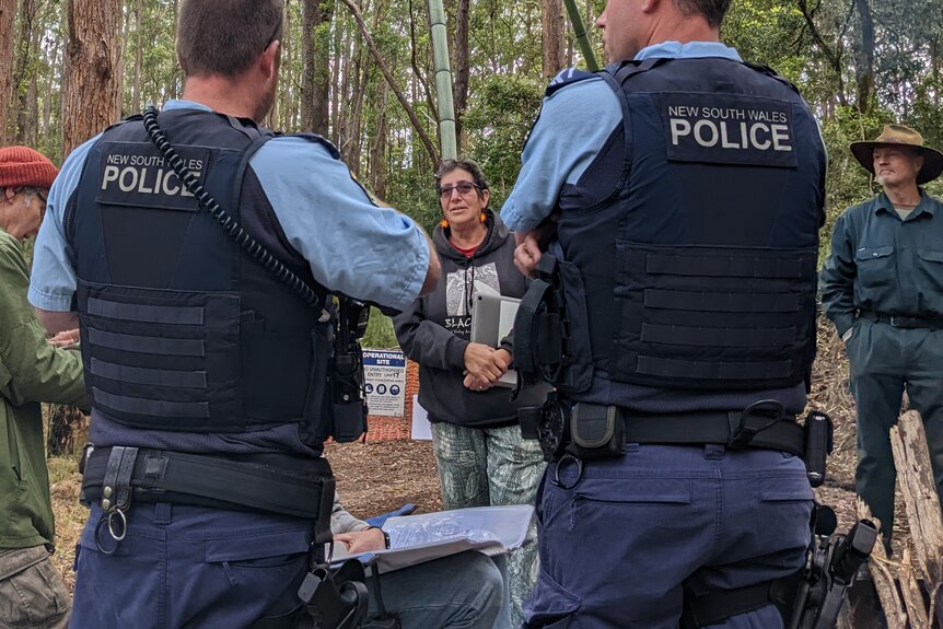 Police with their backs facing the camera, a woman speaking to them with forest in the background