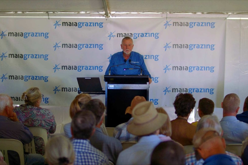 A bearded, middle-aged man in shirt sleeves stands at a dais addressing men and women farmers.