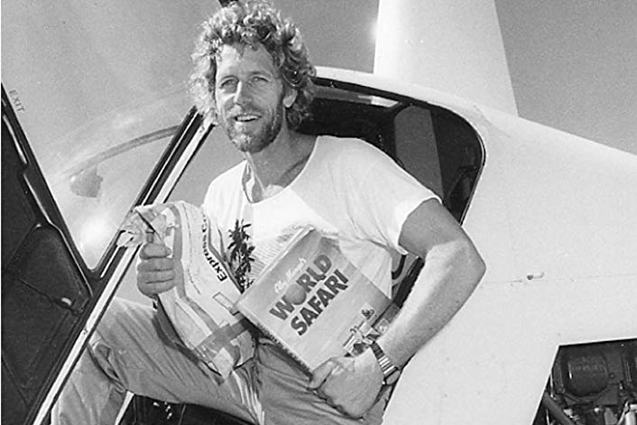 Alby Mangels sits in the door of a helicopter with a copy of his World Safari II under arm.