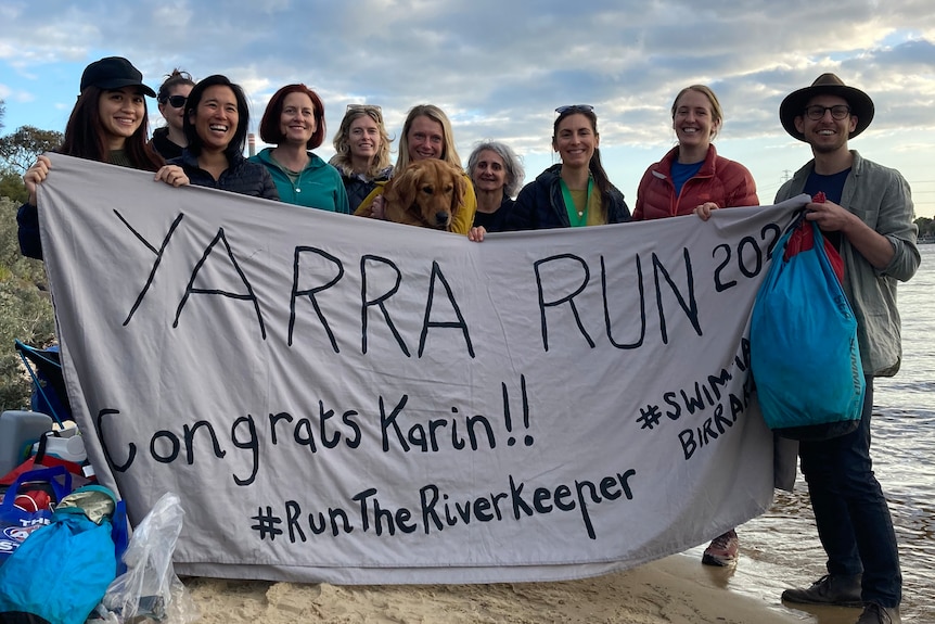 A group of people holding a sign saying 'YARRA RUN 2022 CONGRATS KARIN' under the West Gate Bridge at sunset.