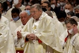 Cardinal George Pell is dressed in a white and gold robe with his hands clasped in front of him.