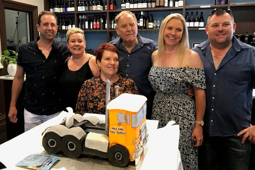 A group of people with a cake that looks like a truck in front of wine bottles on shelves