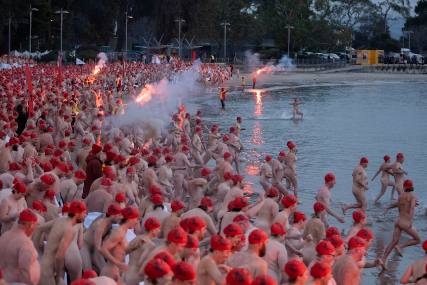 Large crowds of nude people running into the water with red caps on.