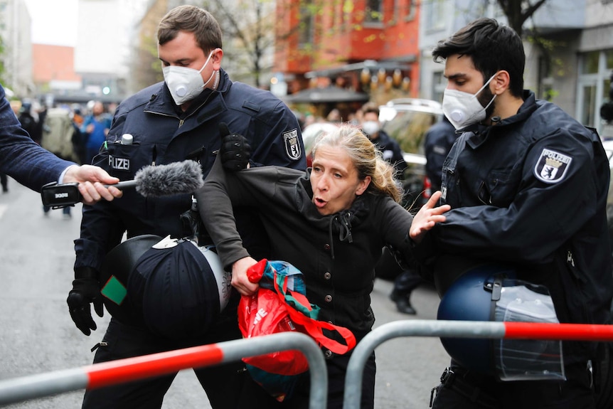 Two police in masks grab a woman by her elbows as she talks into a microphone in front of a barricade.