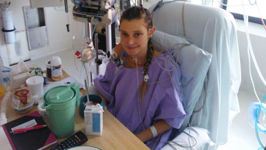 A young woman sits in a hospital bed, surrounded by tubes.