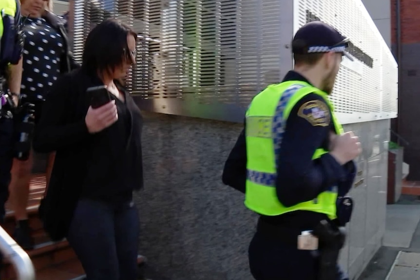 A woman with her face obscured by black hair walks behind a police officer