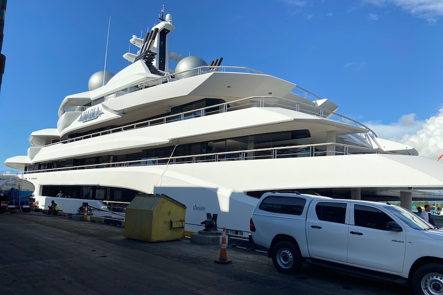 A large superyacht sits next to a pontoon, a host is hooked up from a machine parked up alongside