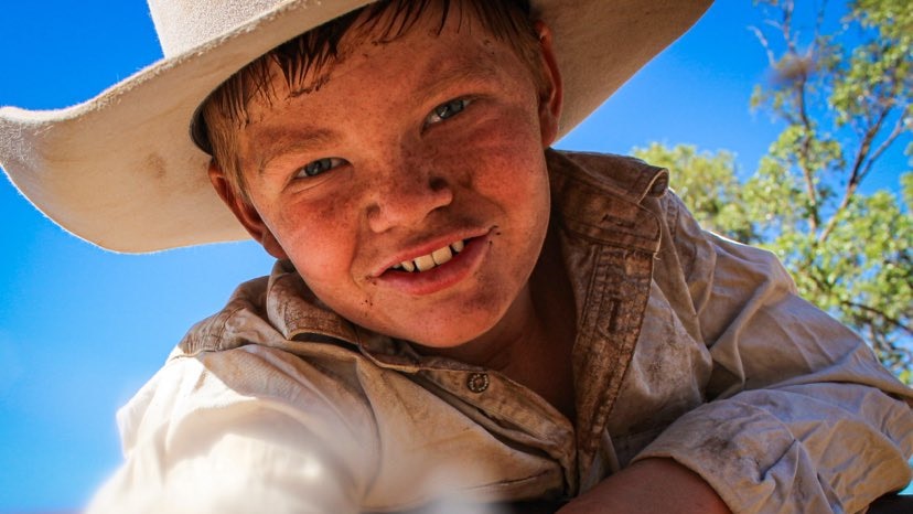 Young boy looking down the barrel of the camera in a cowboy hat, mud smeared on his face.