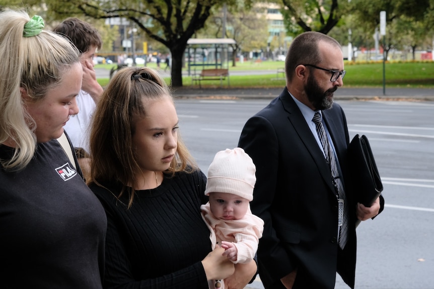 A woman with light brown hair holds a young baby while a blonde woman and a man wearing a suit walk beside them