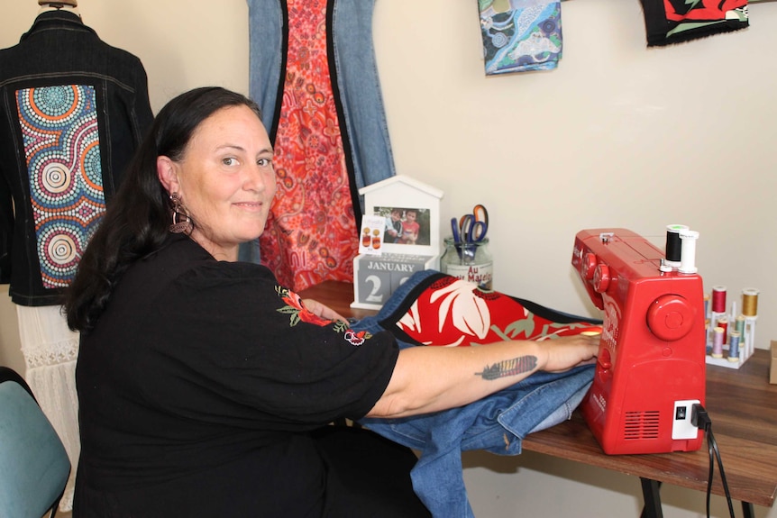 A woman at a sewing machine with clothes hanging up in the background.