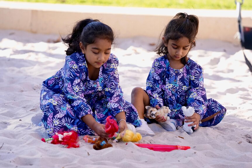 Two young sisters wearing floral dresses have heads down as they sit and play in a sandpit