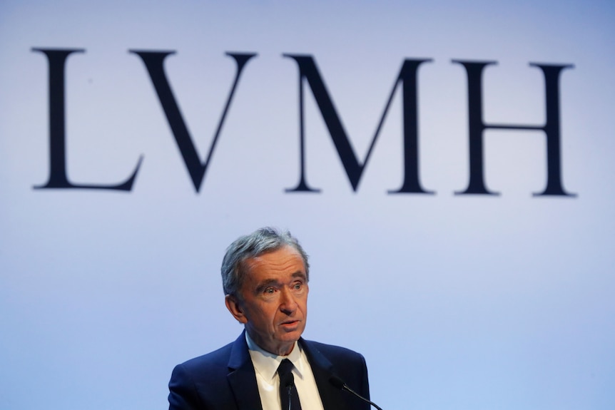 A white man with streaked grey hair dressed in a blue suit stands before the letters LVMH projected behind him
