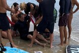 A sitting man in a wetsuit grimaces as his injured arm is raised. He is surrounded by people at a beach.