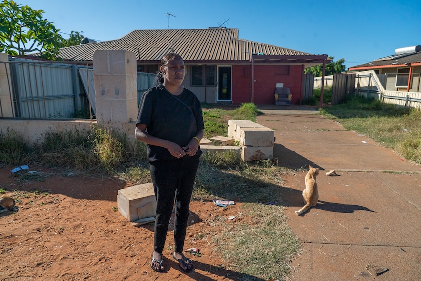 An Indigenous woman stands in front of her house. A brick barrier lies in ruins behind her.