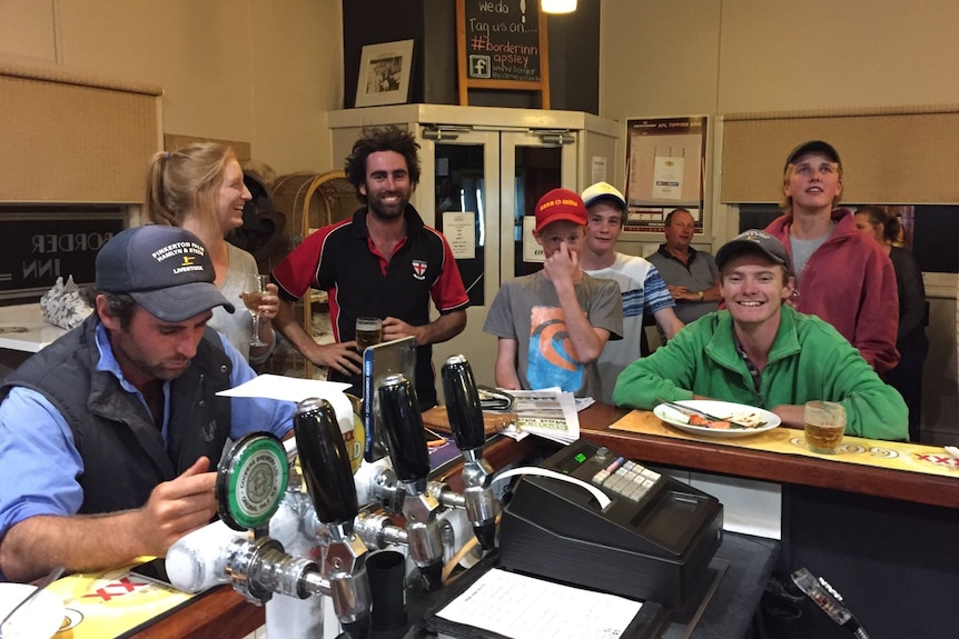 Several men, women and teenagers gather around a smiling pub bar.