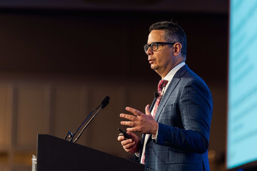 Ben Wyatt stands wearing glasses and a red tie and suit at a business breakfast in Perth.