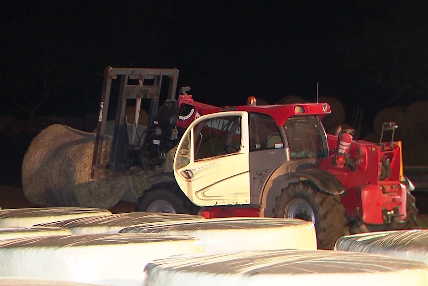 A tractor with round hay bales loaded onto its forks, it is night time