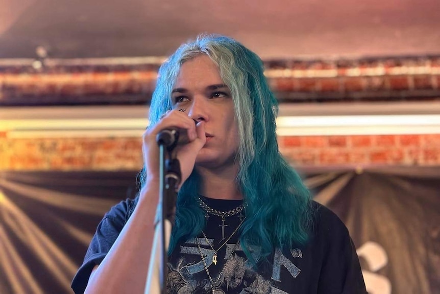 A blue haired young man stands at the microphone and stares out at the crowd. 