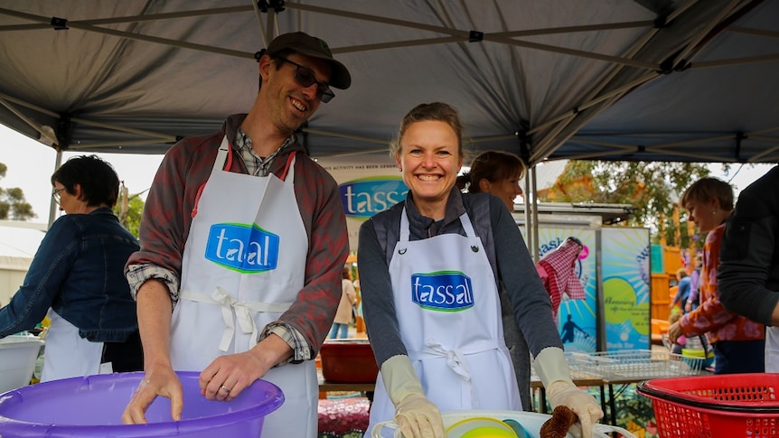 A man and woman smile at the camera while washing dishes in buckets at a school fete.
