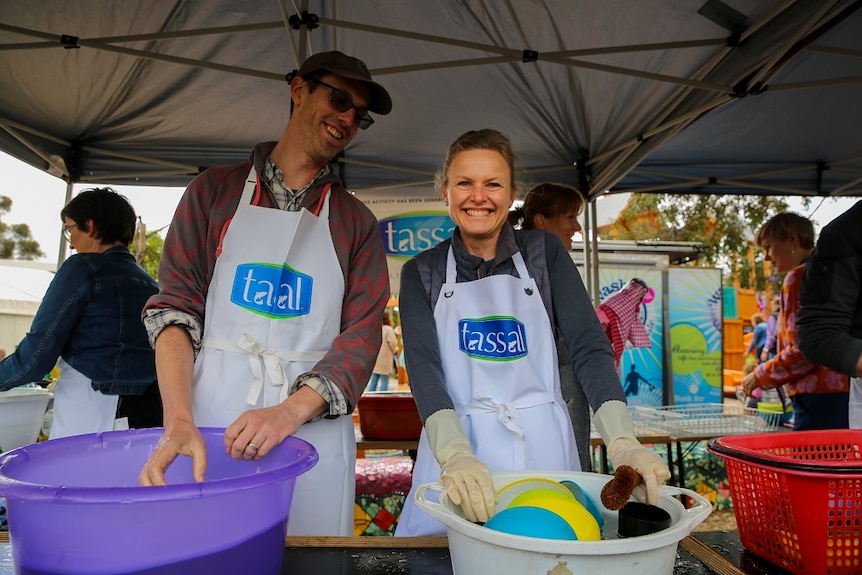 A man and woman smile at the camera while washing dishes in buckets at a school fete.