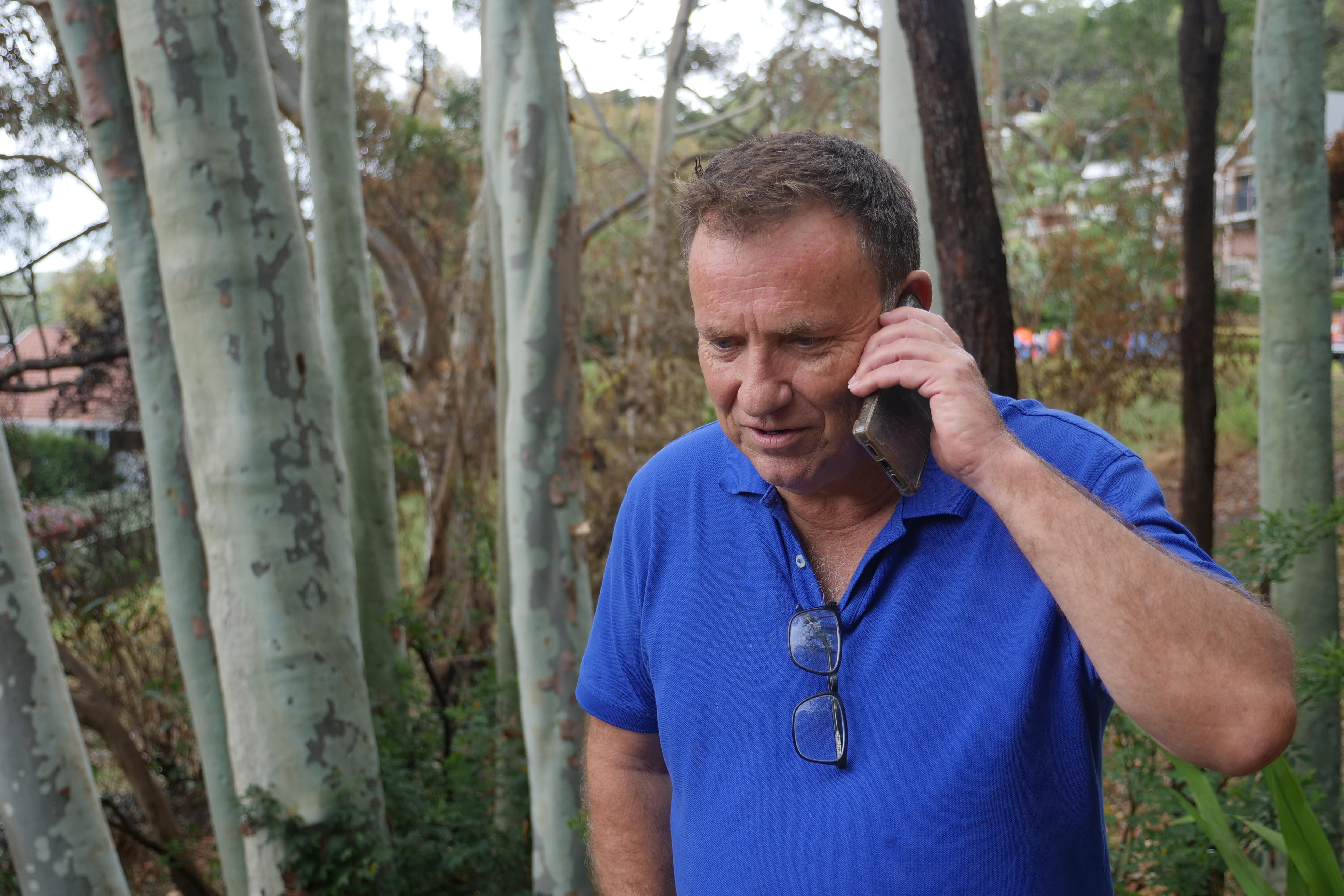 Un hombre de mediana edad, de pelo oscuro, habla por teléfono mientras está parado entre unos árboles en un jardín.