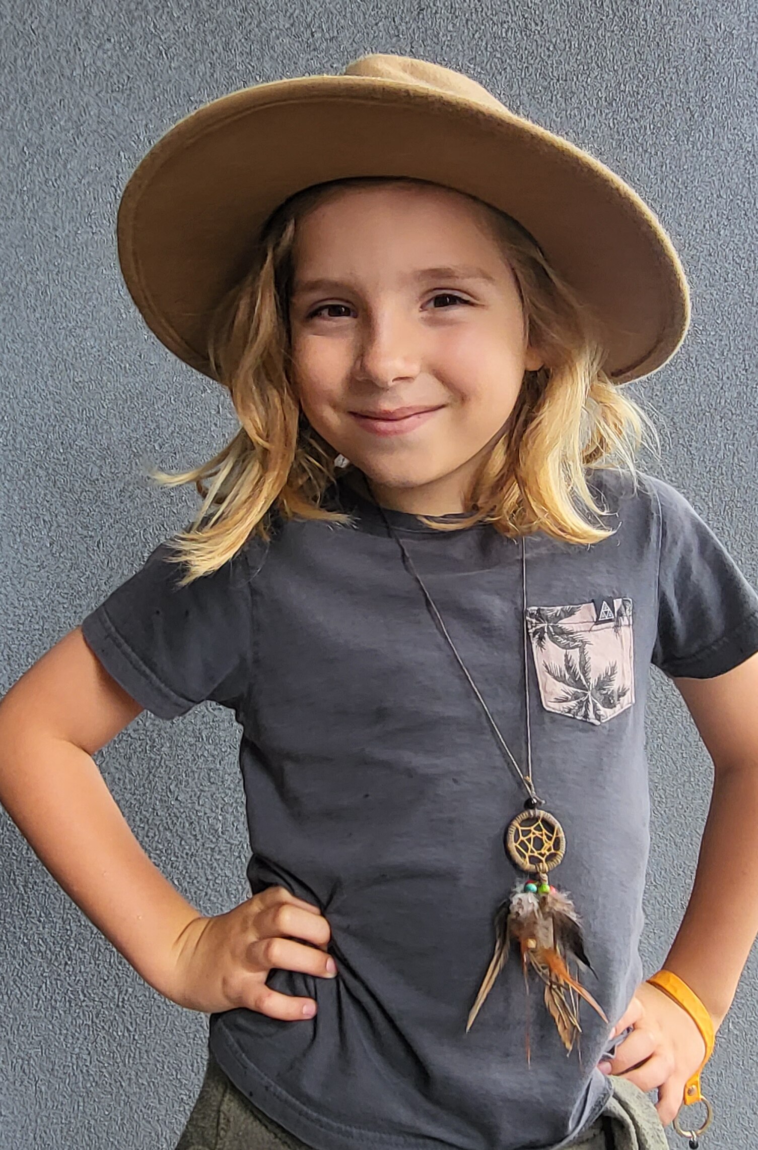 A young boy with long blonde hair and a felt hat smiling