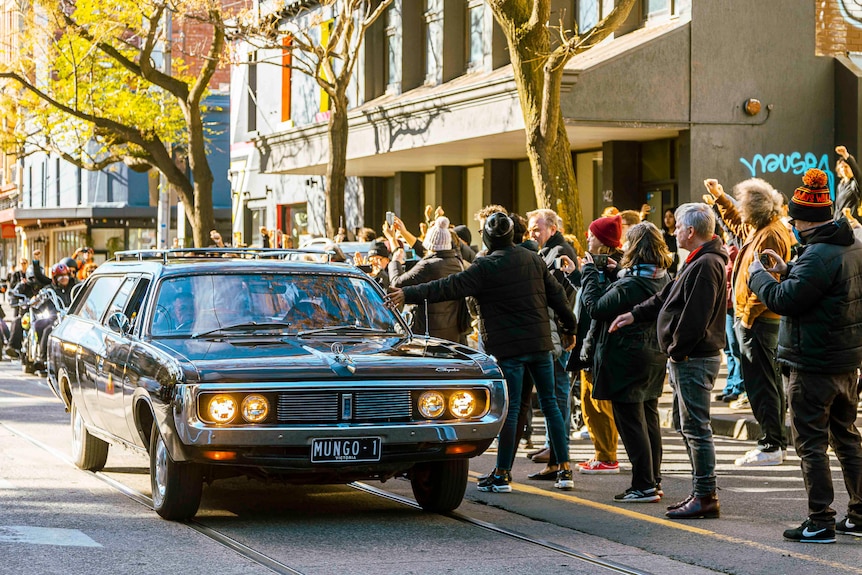 Dozens of people line the footpath, waving to a black funeral car with the license plate 'Mungo 1'.