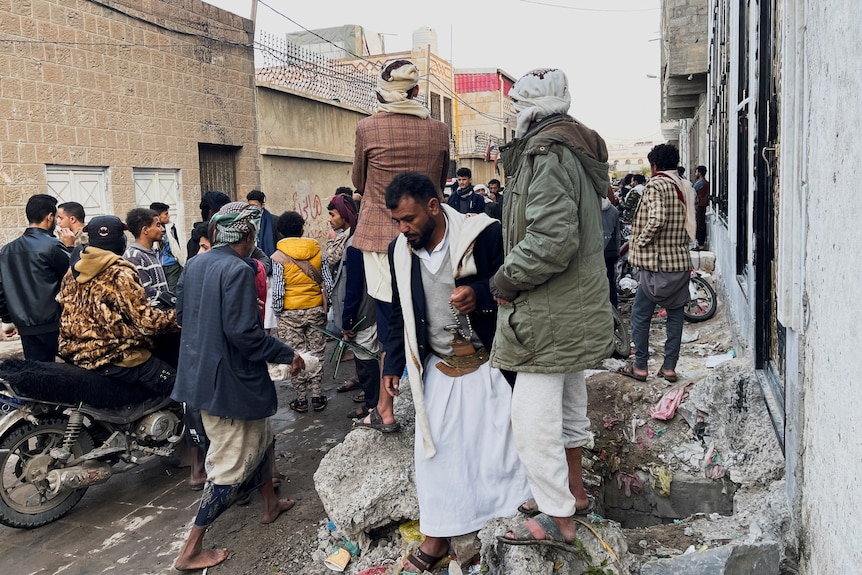 A group of people father outside on a busy street in Yemen, some standing on top of cement littered with rubbish.