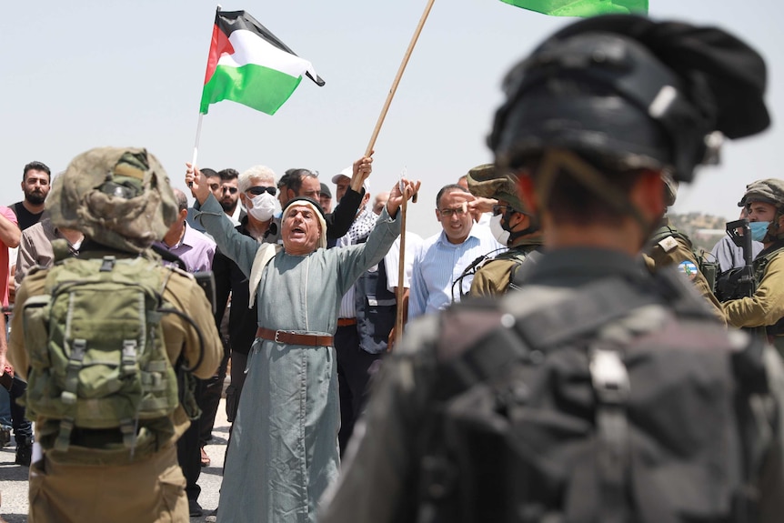 Man holding a Palestinian flag and walking stick stands in front of Israeli soldiers at a check point.