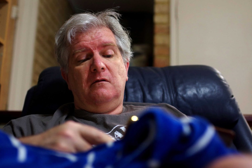 A close-up shot of a man with grey hair lying down on his back with a hand on his chest.