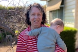 Woman holding an unidentified baby in her front garden. 