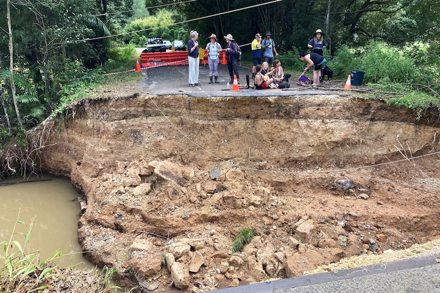Damaged road with people standing on the edge of road and water flowing beneath
